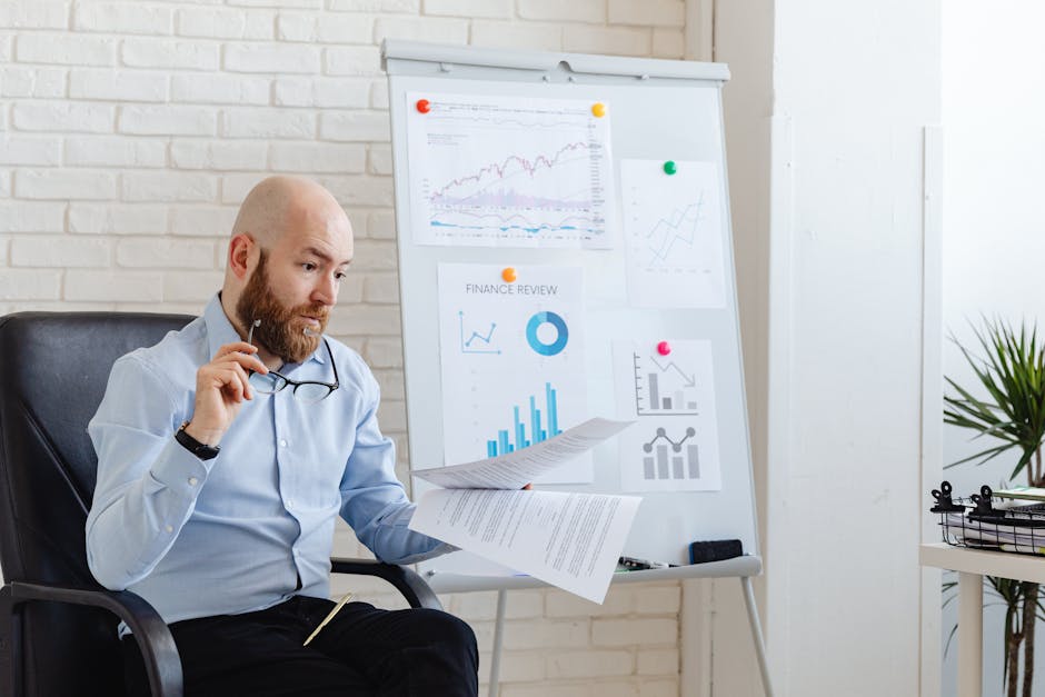 Bald businessman analyzing financial charts in modern office setting with whiteboard and paperwork.